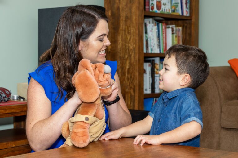 A speech pathologist is holding a dog puppet. She is kneeling in front of a coffee table. A young boy is copying her face. There is a book case behind them.