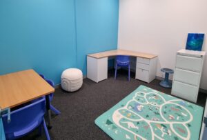 A clinic room, with one blue wall and one white wall. There is a desk in the corner, with a blue chair at it. There is a blue and white playmat on the floor.